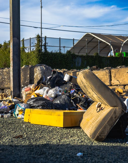 A collection of discarded rubbish including black plastic trash bags, a yellow plastic storage box, a large, dirty beige car seat with fabric upholstery, and scattered debris lying on a gravel surface. The rubbish appears to be gathered for collection or removal, with some items leaning against a stone wall made of irregularly shaped, weathered stones. Behind the wall, there is a fenced area with greenery and trees, and beyond that, a large tent-like structure with a curved roof is visible outdoors. The scene is illuminated by natural daylight, casting shadows and highlighting the textures of the waste and surrounding environment, which represents a typical scene for private rubbish removal services in an outdoor setting.