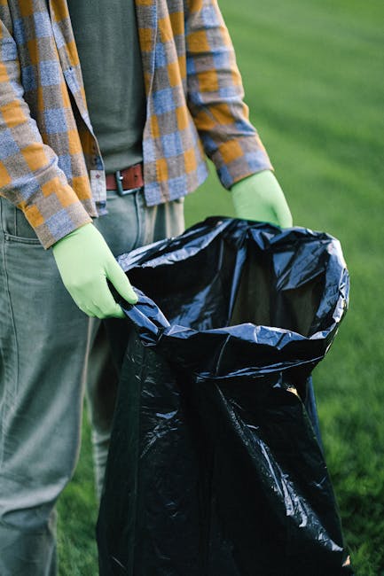 A person wearing a green long-sleeved shirt, a checkered yellow and grey jacket, and grey trousers is holding open a black plastic rubbish bag by its edges, preparing to dispose of waste. The individual is wearing light green gloves for handling waste materials. The scene is set outdoors on a grassy area with a blurred green background, possibly a park or garden, suggesting a cleanup activity or waste collection in a natural environment. The black rubbish bag is made of shiny plastic, appearing partially filled and ready for waste collection or removal. The person's posture indicates they are in the process of either lining the bag or readying it for rubbish disposal, aligning with services related to rubbish removal or waste management, such as those offered by Rubbish Removal Bow, in the context of private waste handling outside of official council collection. The overall scene emphasizes environmentally conscious waste disposal in a tidy and organized manner.