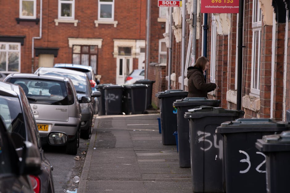 A residential street scene showing a row of black wheelie bins lined up on the pavement outside brick terraced houses, which have white-framed windows and stone window sills. The bins are positioned adjacent to the houses, with their lids closed, and some display white graffiti or markings. In the background, there are additional bins and parked cars, including a silver hatchback, with the street extending into the distance. A woman dressed in a brown jacket and hood is seen standing next to one of the bins, appearing to be handling rubbish or preparing to dispose of waste. The scene is illuminated by natural daylight, highlighting the textured brickwork of the buildings and the urban environment, which is typical for private waste collection areas, consistent with independent rubbish removal services like those offered by Rubbish Removal Bow. The overall setting reflects a typical domestic refuse collection point, focused on waste management through private waste handling methods, with emphasis on the arrangement and functioning of waste bins in an urban residential area.