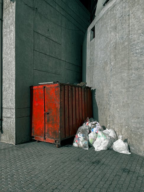 A large, rectangular metal waste skip painted in red, with visible signs of rust and wear, is positioned against a dark green wall on the left and a light gray concrete wall on the right, located on a paved urban outdoor area. The skip is filled with loose rubbish and trash bags, some of which are placed on the ground nearby. The trash bags are made of clear and white plastic, and contain various discarded items, including paper, plastic bottles, and other household waste. The surrounding environment features a textured concrete quadrant wall and a dark asphalt pavement with a patterned surface. Better lighting highlights the textures of the rusted metal and the smooth plastic bags, capturing the scene of an outdoor rubbish collection point that could be serviced by independent rubbish removal providers like Rubbish Removal Bow, offering an alternative to council collections for Bow residents seeking private waste disposal options.