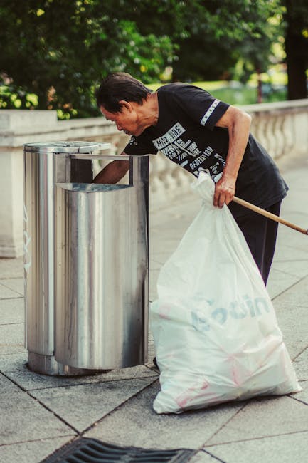 A middle-aged man with dark hair, wearing a black t-shirt with white text on the front and dark trousers, is actively discarding rubbish into an open-topped stainless steel public bin situated on a paving area. The bin has a rounded, modern design with a smooth, reflective surface, and is positioned near a low stone wall with a decorative railing. Behind the man, a lush green environment with trees and grass is visible, indicating an outdoor park or public space setting. The man holds a large white plastic bag filled with rubbish in his left hand, which appears to contain various waste items, and uses his right hand to place debris into the bin. The scene is well-lit with natural daylight, emphasizing the clean, organized environment suitable for neighbourhood rubbish collection or enhanced waste management solutions like private disposal services, as offered by Rubbish Removal Bow.