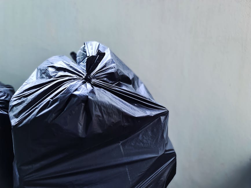 A large, rectangular metal waste skip painted in red, with visible signs of rust and wear, is positioned against a dark green wall on the left and a light gray concrete wall on the right, located on a paved urban outdoor area. The skip is filled with loose rubbish and trash bags, some of which are placed on the ground nearby. The trash bags are made of clear and white plastic, and contain various discarded items, including paper, plastic bottles, and other household waste. The surrounding environment features a textured concrete quadrant wall and a dark asphalt pavement with a patterned surface. Better lighting highlights the textures of the rusted metal and the smooth plastic bags, capturing the scene of an outdoor rubbish collection point that could be serviced by independent rubbish removal providers like Rubbish Removal Bow, offering an alternative to council collections for Bow residents seeking private waste disposal options.